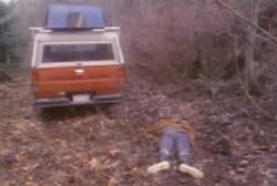 A body is laying face down on the ground in a forest, there is a truck parked nearby.