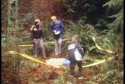 Three police officers stands next to the corpse, covered by a white sheet. There is crime scene tape circling the area.