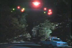 A police cruiser stopped in the middle of a road facing an unidentified flying object in the sky