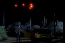 A man outside his car looking up at a tight arrangment of red lights in the night sky over the desert