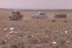 Three cars driving towards metal debris scattered aroud a small area in the desert
