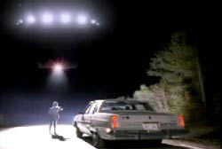 Betty Cash standing next to her car on a deserted road looking up at a flying disk in the sky with many lights (UFO)