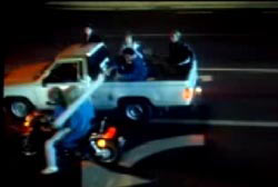 While driving parallel to each other a man in the bed of a pickup truck strikes the head of a woman on the back of a motorcycle